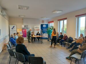 Sala pełna ludzi siedzących na krzesłach, kobieta w żółtej koszuli stoi w centrum.