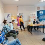 Kobieta w żółtej koszuli występuje przed osobami siedzącymi na krzesłach.