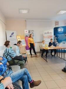 Kobieta w żółtej koszuli występuje przed osobami siedzącymi na krzesłach.