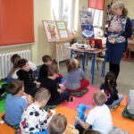 Grupka siedzących dzieci słucha stojącej blondwłosej kobiety.