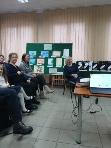 Kilka osób w różnym wieku siedzi na krzesłach, w tle zielona tablica z pracami plastycznymi, oraz stół z laptopem.