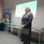 Blondwłosa kobieta w długiej sukience stoi przed gablotami i prezentacją.