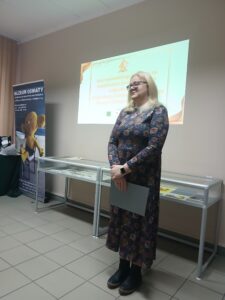 Blondwłosa kobieta w długiej sukience stoi przed gablotami i prezentacją.