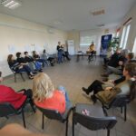 W dużej sali osoby siedzą na krzesłach przy ścianach. W centrum stoi kobieta w żółtej koszuli.