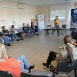 W dużej sali osoby siedzą na krzesłach przy ścianach. W centrum stoi kobieta w żółtej koszuli.