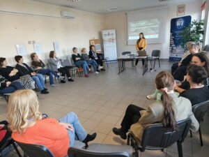 W dużej sali osoby siedzą na krzesłach przy ścianach. W centrum stoi kobieta w żółtej koszuli.