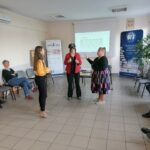 Kobieta w żółtej koszuli tłumaczy coś dwóm kobietom: w czerwonej marynarce i czarnej sukience.