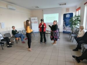 Kobieta w żółtej koszuli tłumaczy coś dwóm kobietom: w czerwonej marynarce i czarnej sukience.