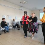 Kobieta w czerni prowadzi pod ramię kobietę w czerwonej marynarce z opaską na oczach.