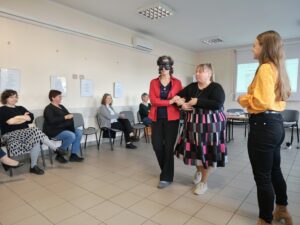 Kobieta w czerni prowadzi pod ramię kobietę w czerwonej marynarce z opaską na oczach.