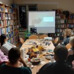 Osoby siedzące przy stolikach oglądają prezentację na ekranie.