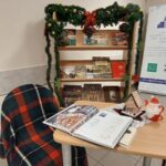 Regał z książkami ozdobiony girlandą, przed nim krzesło z kocem w kratę i stolik z książką.