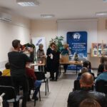 Na środku sali stoją 3 kobiety i wręczają prezent dziewczynce. W tle siedzą ludzi na krzesłach. Mężczyzna robi zdjęcie aparatem.