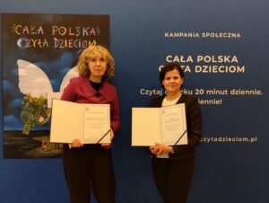 Blondynka i brunetka pozują na tle niebieskiej ścianki z dyplomami w dłoniach.