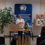 Sala audytoryjna, siedzi przodem do kobiety w bieli stojącej na środku sali.