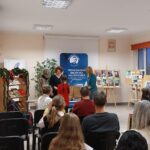 Na środku sali stoją 3 kobiety i wręczają prezent dziewczynce. W tle siedzą ludzi na krzesłach.