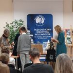 Kobieta w zielonej sukience trzyma czarną torbę, chłopiec w szarym swetrze stoi przodem do dwóch innych kobiet. Z tylu siedzący ludzie.