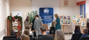Kobieta w zielonej sukience trzyma czarną torbę, chłopiec w szarym swetrze stoi przodem do dwóch innych kobiet. Z tylu siedzący ludzie.