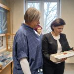 Dwie kobiety w sali muzealnej pochylają się nad grafiką w dłoni jednej z nich.