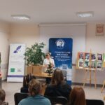 Sala audytoryjna, siedzi przodem do kobiety w bieli stojącej na środku sali.