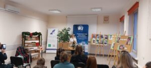 Sala audytoryjna, siedzi przodem do kobiety w bieli stojącej na środku sali.
