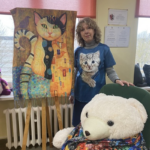 Blondwłosa kobieta pozuje przy dużym pluszaku i portrecie kota.