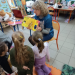 Blondwłosa kobieta pokazuje książkę dzieciom.