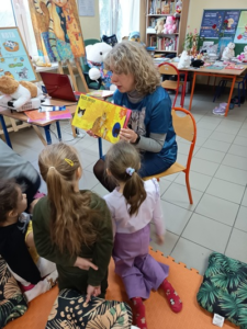 Blondwłosa kobieta pokazuje książkę dzieciom.