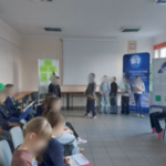 Na przodzie sali stoi kilku uczniów. Część siedzi na krzesłach.