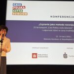 Kobieta przed mikrofonem. W tle duży ekran z nazwą konferencji.