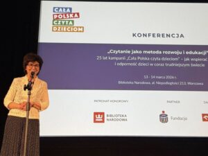 Kobieta przed mikrofonem. W tle duży ekran z nazwą konferencji.