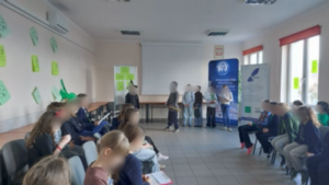 Na przodzie sali stoi kilku uczniów. Część siedzi na krzesłach.