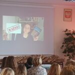 Sala audytoryjna pełna słuchaczy, na rozwiniętym ekranie kobieta w okularach trzyma niebieską książkę.