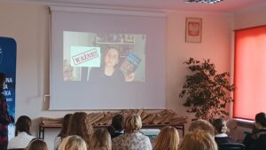 Sala audytoryjna pełna słuchaczy, na rozwiniętym ekranie kobieta w okularach trzyma niebieską książkę.