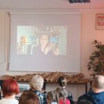 Sala audytoryjna pełna słuchaczy, na rozwiniętym ekranie kobieta w okularach trzyma niebieską książkę.