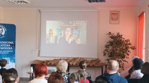 Sala audytoryjna pełna słuchaczy, na rozwiniętym ekranie kobieta w okularach trzyma niebieską książkę.