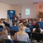 Sala audytoryjna pełna słuchaczy, dwie kobiety wręczają nagrodę trzeciej.