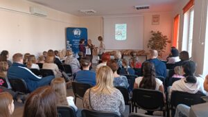 Sala audytoryjna pełna słuchaczy, dwie kobiety wręczają nagrodę trzeciej.