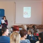 Sala audytoryjna pełna słuchaczy, jako mówca stoi kobieta w kwiecistej sukience.