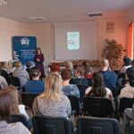 Sala audytoryjna pełna słuchaczy, jako mówca stoi kobieta w kwiecistej sukience.