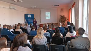 Sala audytoryjna pełna słuchaczy, jako mówca stoi kobieta w kwiecistej sukience.