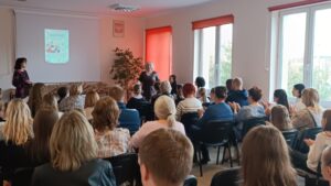 Sala audytoryjna pełna słuchaczy, jako mówca stoi kobieta w brązowej sukience.