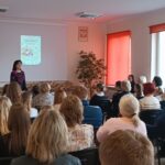 Sala audytoryjna pełna słuchaczy, jako mówca stoi kobieta w kwiecistej sukience.