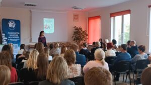 Sala audytoryjna pełna słuchaczy, jako mówca stoi kobieta w kwiecistej sukience.