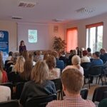 Sala audytoryjna pełna słuchaczy, jako mówca stoi kobieta w kwiecistej sukience.