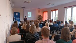 Sala audytoryjna pełna słuchaczy, jako mówca stoi kobieta w kwiecistej sukience.