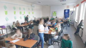 Sala zajęciowa. Dzieci siedzą w kilku grupach przy stolikach.