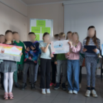 Kilkoro dzieci stoi. Część z nich trzyma prace plastyczne.