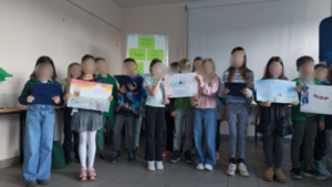 Kilkoro dzieci stoi. Część z nich trzyma prace plastyczne.