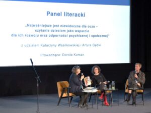 Dwoje panelistów i prowadząca dyskusję przed dużym ekranem.